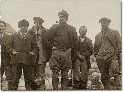 Photo of six men. Some are smiling. One man is smoking a pipe. They are wearing coats and boats and standing outside.