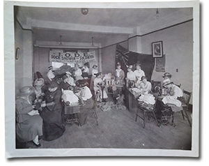 historical photo of several women in a room with a banner in the background that reads “I.O.D.E Sock Shower for Soldiers Headquarters, 3rd floor, free wool to knitters.” Many of the women are kitting or operating sewing machines, while others pour tea.