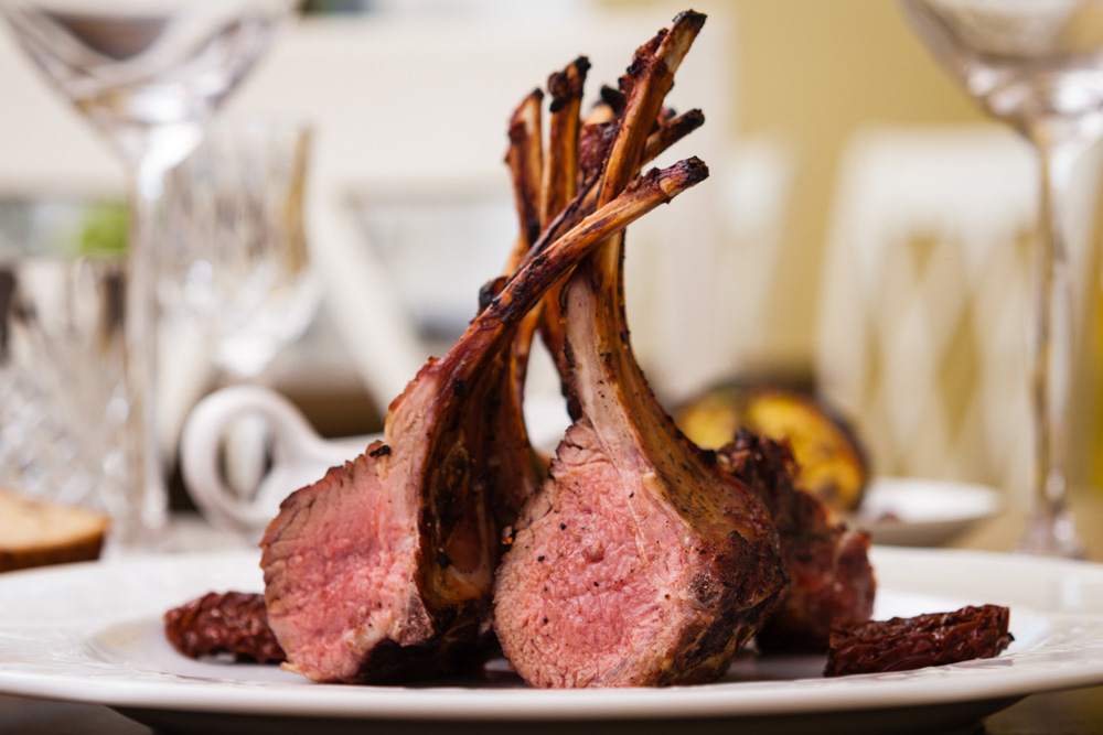Picture of two racks of lamb cooked to medium rare served on a dish with crystal ware wine glasses in the background