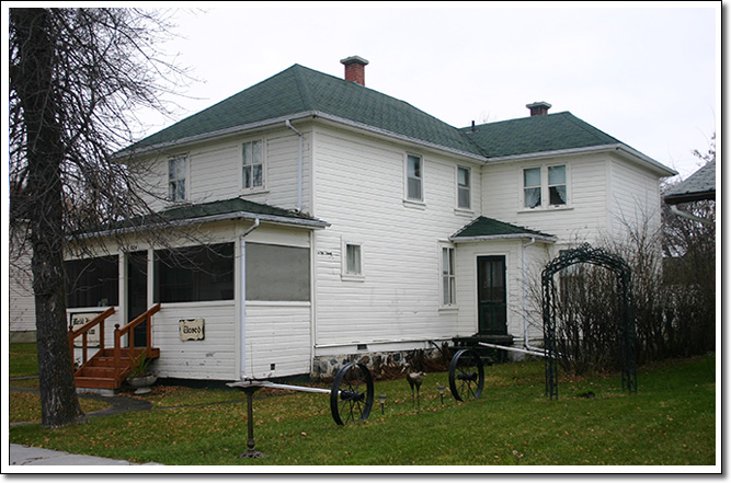 Une maison à ossature de deux étages peinte en blanc sur un plan en forme de L, avec un toit en croupe et une véranda grillagée