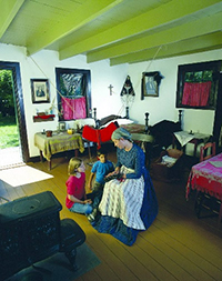 Interior of old house with a female interpreter dressed in period costume showing a Métis sash to two children