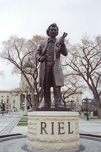 Bronze statue of the full figure of Louis Riel holding a document in his fist. Figure is on a pedestal that reads RIEL.